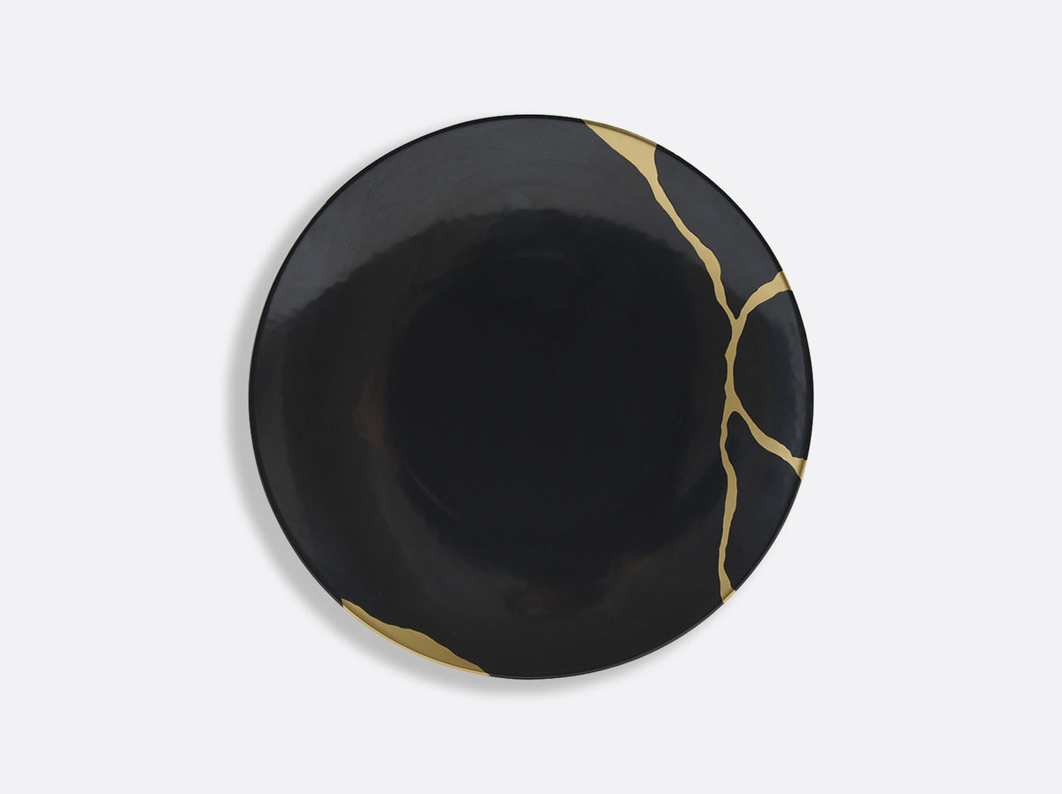 The Bernardaud Kintsugi Dinnerware features a round, black ceramic plate accented with gold lines inspired by the Japanese art of repairing with gold, creating a striking contrast on its glossy surface.