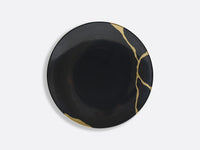 The Bernardaud Kintsugi Dinnerware features a round, black ceramic plate accented with gold lines inspired by the Japanese art of repairing with gold, creating a striking contrast on its glossy surface.