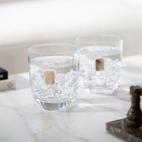 Two Elevo DOF Glasses Smoke by Anna New York, filled with ice and water, sit on a white marble surface in bright natural light. Nearby are a small chess piece and part of a rectangular object. Set includes 2 lead-free crystal glasses.