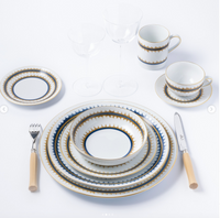 A refined table setting with Marie Daâge Rafia Stripes patterned plates, bowls, cups, and saucers—hand-painted Limoges porcelain—paired with wine glasses and wooden-handle cutlery on a white background.