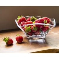 A Simon Pearce Woodbury Bowl, crafted from clear handblown glass, holds fresh strawberries on a wooden surface, with two strawberries placed beside it.