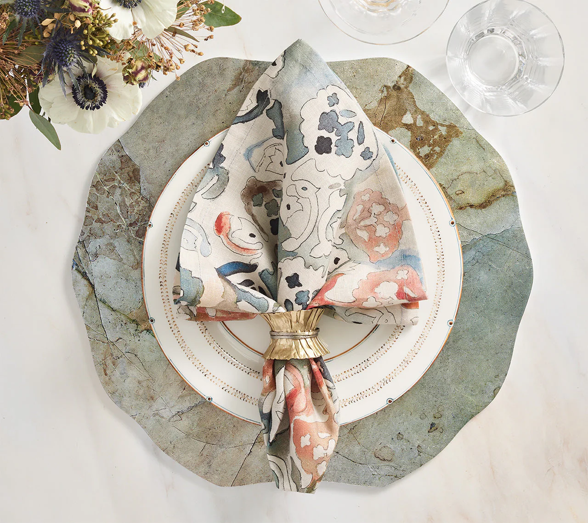 A modern table setting showcases a scalloped stone-patterned placemat, white plate with gold trim, floral napkin with Kim Seybert’s Clinched Napkin Ring in Gold & Silver (set of 4), elegant glassware on marble, and a nearby flower arrangement.