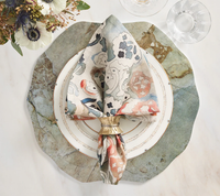 A modern table setting showcases a scalloped stone-patterned placemat, white plate with gold trim, floral napkin with Kim Seybert’s Clinched Napkin Ring in Gold & Silver (set of 4), elegant glassware on marble, and a nearby flower arrangement.