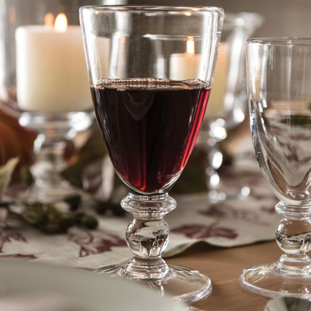 A Simon Pearce Hartland Glass, made from lead-free glass, is filled with red wine on a dining table with another empty Hartland Glass, lit candles, and a stylish patterned runner for an elegant setting.