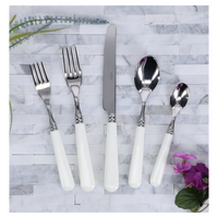 A Capdeco Altea Flatware 5 Piece Setting—stainless steel with white handles—is arranged on a white and gray tiled surface, accented by green leaves and purple flowers. Made in France.