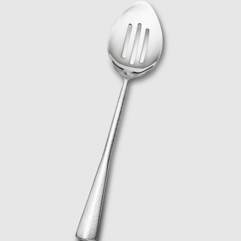 The Mary Jurek Alta Slotted Serving Spoon is featured on a light gray background, showcasing its textured handle and elegant, premium stainless steel design.