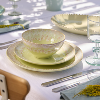 Costa Nova’s Taoromina Dinnerware Yellow—a ceramic bowl, salad plate, and dinner plate with decorative edges—are stacked on a white tablecloth, surrounded by cutlery, a blue napkin, glassware, and yellow flowers in soft sunlight.