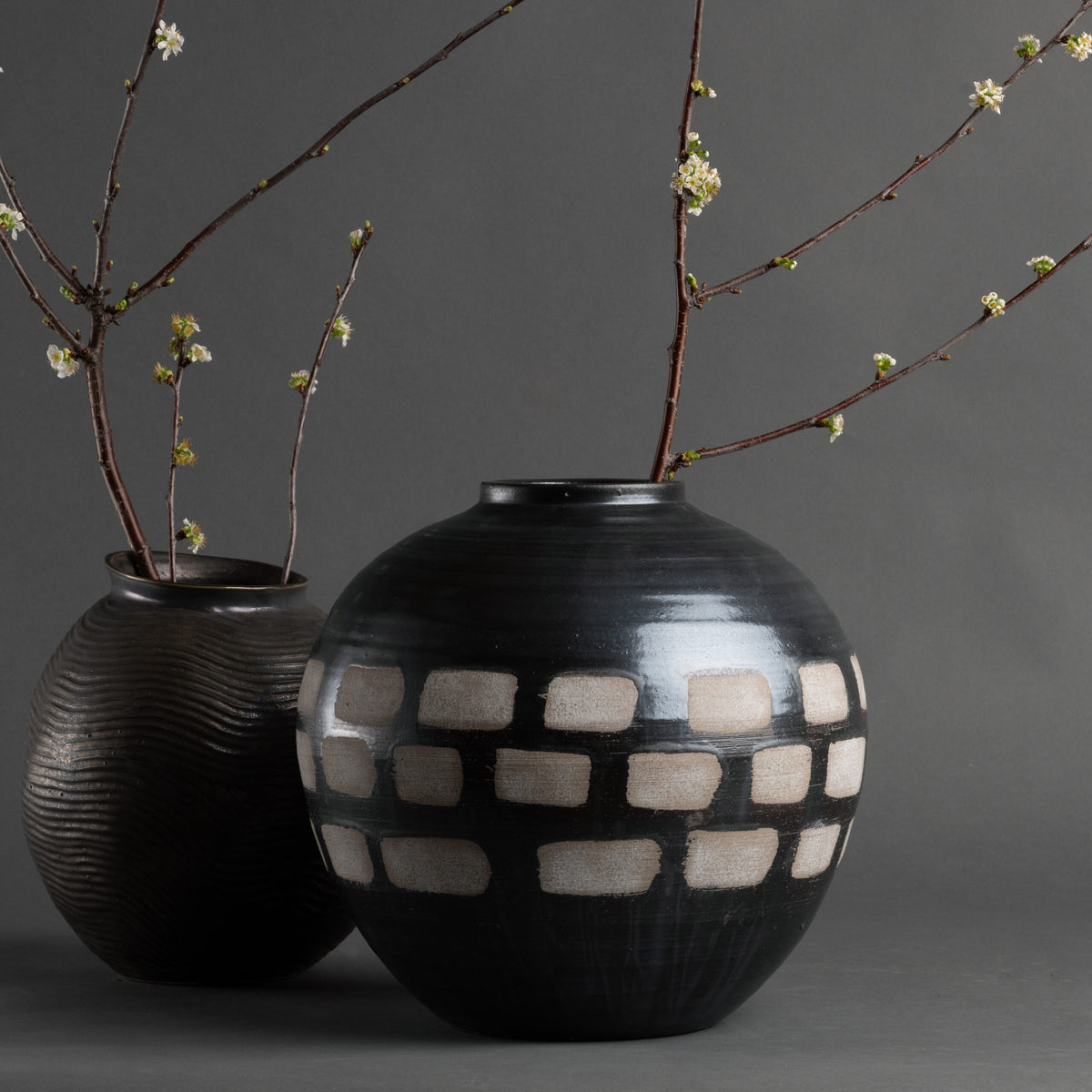 The Jude Vase by FlowDecor Inc.—two black and brown handmade ceramics, one round with a white geometric pattern and one ribbed—holds budding white floral branches, bringing organic modern decor to a simple gray backdrop.