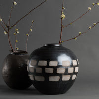 The Jude Vase by FlowDecor Inc.—two black and brown handmade ceramics, one round with a white geometric pattern and one ribbed—holds budding white floral branches, bringing organic modern decor to a simple gray backdrop.
