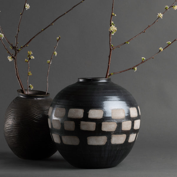 The Jude Vase by FlowDecor Inc.—two black and brown handmade ceramics, one round with a white geometric pattern and one ribbed—holds budding white floral branches, bringing organic modern decor to a simple gray backdrop.