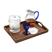 A wicker tray features the Caspari Acrylic Ice Bucket - Clear with ice, a glass pitcher with a blue handle filled with a light brown drink, a blue glass, and stacked white napkins—ideal for stylish barware service.