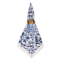 The Iberian Napkin Indigo by Juliska, a set of four white cloth napkins with blue floral patterns inspired by cobalt tiles, is neatly folded and secured with a wooden napkin ring.