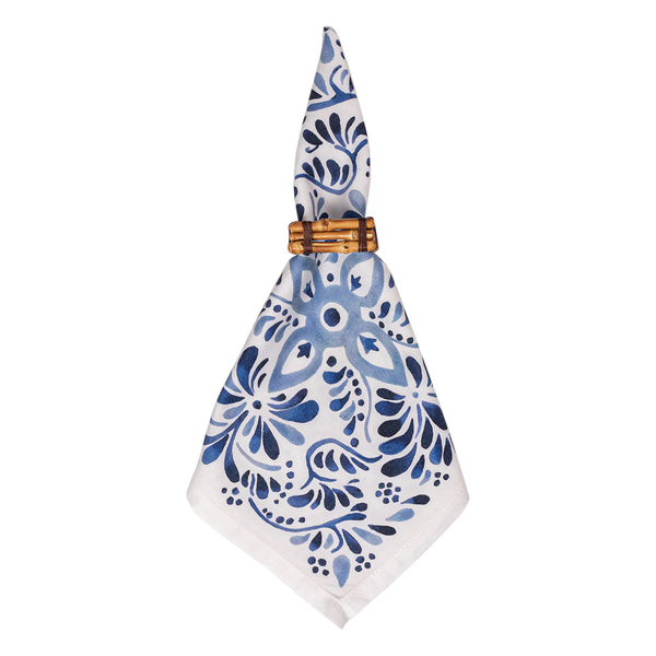 The Iberian Napkin Indigo by Juliska, a set of four white cloth napkins with blue floral patterns inspired by cobalt tiles, is neatly folded and secured with a wooden napkin ring.