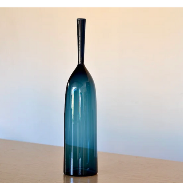 The Angelic Bottle - Small - by Cariati, a tall, slender dark blue glass vase with a narrow neck and wide bulbous base, stands on a wooden surface against a light background.