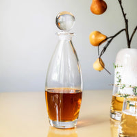 The Addison Decanter by Simon Pearce, filled with amber liquid, sits on a light surface. In the background, a vase with branches bearing yellow pears adds an elegant touch to the barware display.
