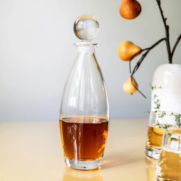 The Addison Decanter by Simon Pearce, filled with amber liquid, sits on a light surface. In the background, a vase with branches bearing yellow pears adds an elegant touch to the barware display.