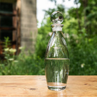The Addison Decanter by Simon Pearce, featuring a round stopper, sits half-filled with water on a wooden table outdoors, set against green foliage and a blurred building in the background.