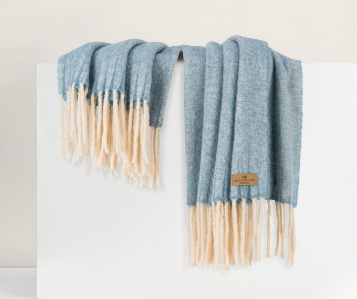 The Andes Throw by Lands Downunder, in light blue alpaca wool with a thick cream fringe and a small brown label, is draped over a white surface.