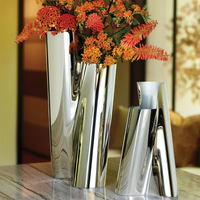 Two modern, reflective silver vases sit on a marble surface. One vase—the Lituus Vase by Georg Jensen—holds red and orange flowers, while the other is empty. Both hand-polished stainless steel vases feature an abstract, folded design. Warm, blurred background.