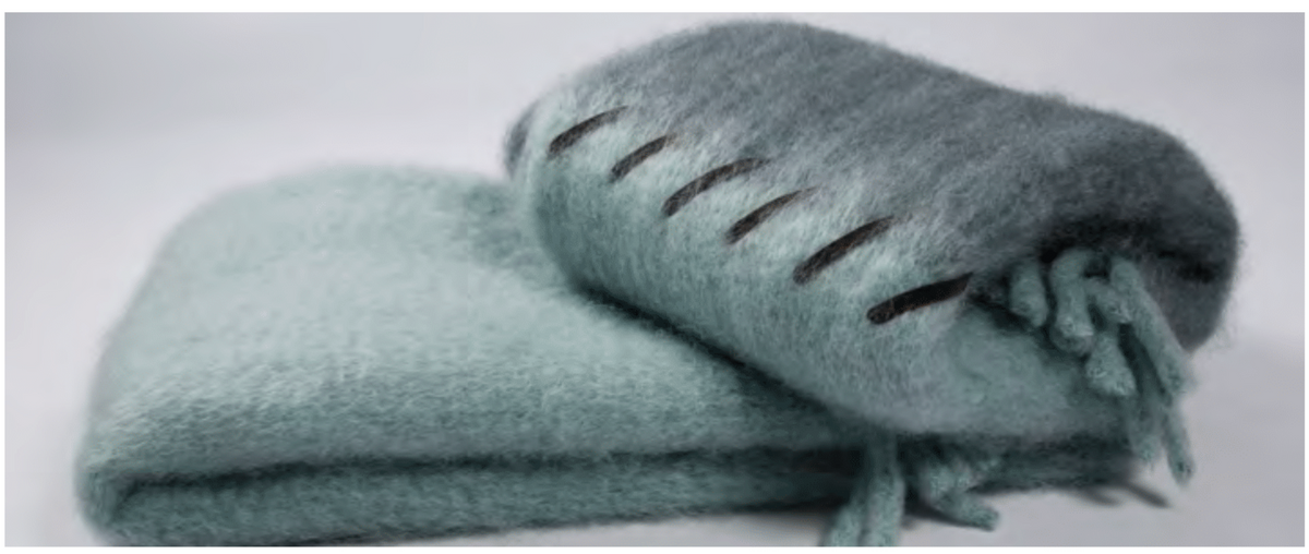 Two soft, fluffy light blue-green Stitching Throw blankets by Mantas Ezcaray—one folded, one rolled with visible fringe and dark stitching—bring handmade luxury style, each placed on a plain white background.