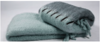 Two soft, fluffy light blue-green Stitching Throw blankets by Mantas Ezcaray—one folded, one rolled with visible fringe and dark stitching—bring handmade luxury style, each placed on a plain white background.