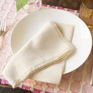 A white plate holds a neatly folded Dove & Donkey Hemstitch Classic Napkin Cloud (Set of 4), set on a pink textured placemat beside a fork, glass, and small vase with a green leaf.