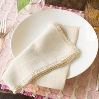 A white plate holds a neatly folded Dove & Donkey Hemstitch Classic Napkin Cloud (Set of 4), set on a pink textured placemat beside a fork, glass, and small vase with a green leaf.