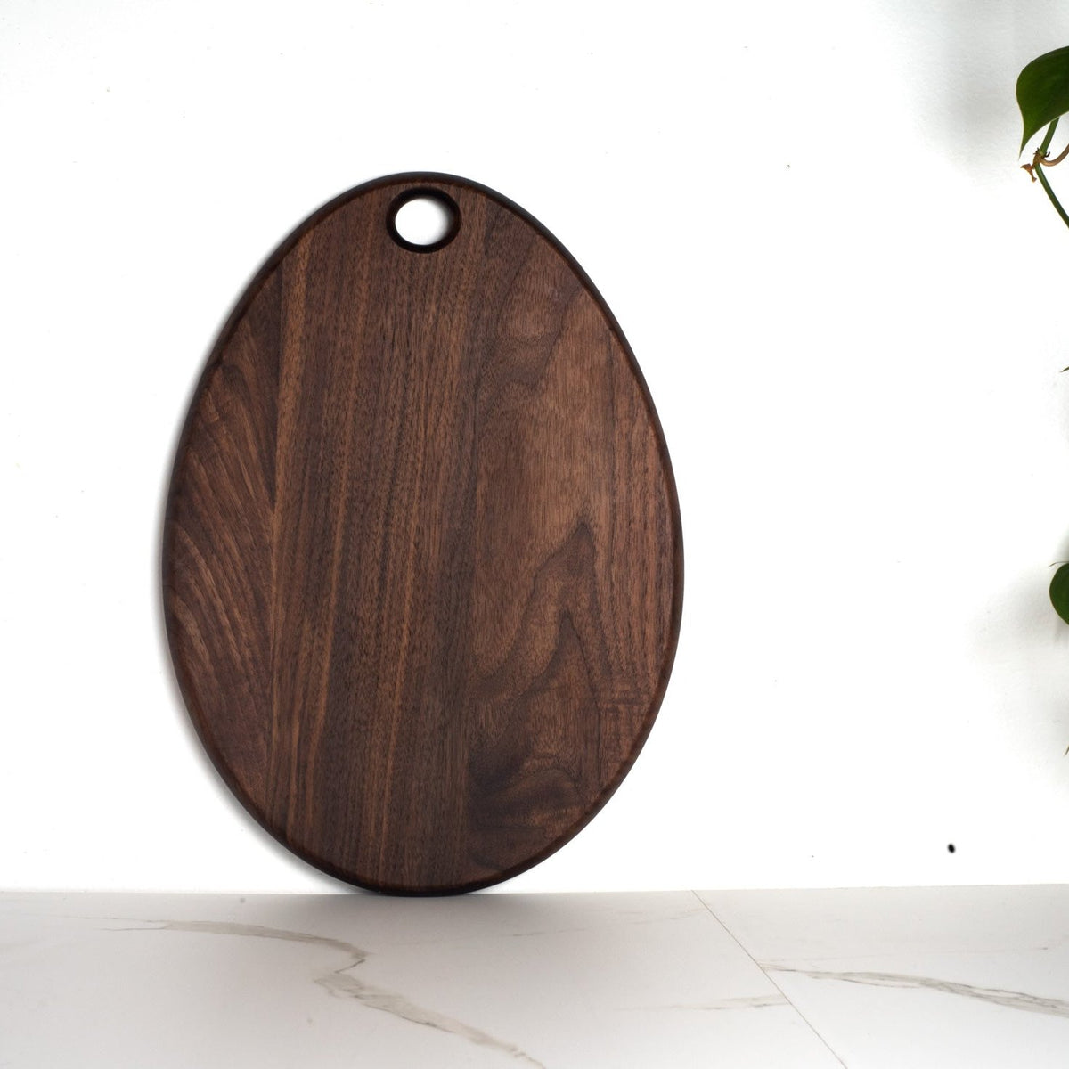 Khem Oval Serving Board Walnut, an oval wooden serving board perfect as a compact cutting board, featuring a circular hanging hole at the top.