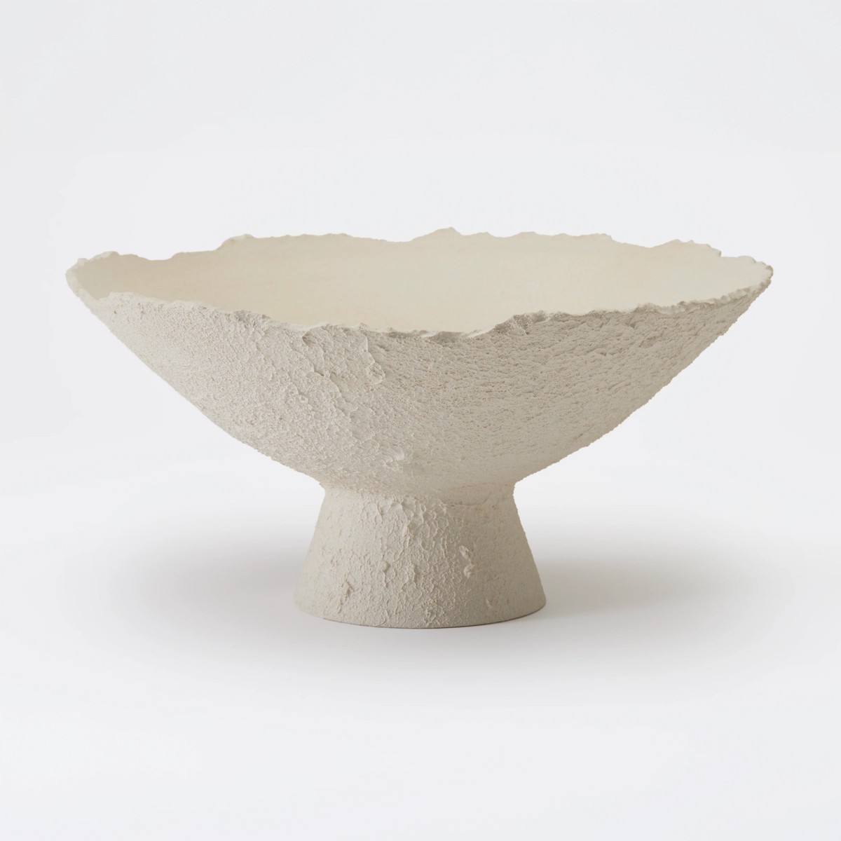 The Joelle Bowl by Dumae features a Sierra-textured, off-white finish with rough edges and a wide, shallow form. Its short cylindrical pedestal base highlights artisanal craftsmanship against a plain white background.