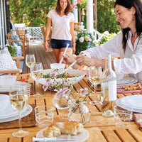 Two women dine outdoors on a wooden patio. One serves salad onto Mariposa Swirl Alabaster Dinnerware White Set of 4 plates at a set table with wine glasses and flowers, as the other approaches, smiling in the bright green setting.