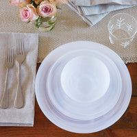 A table is set with Mariposa Swirl Alabaster Dinnerware White Set of 4, a bowl on a woven placemat, two forks on a gray napkin, a snowflake-patterned dishwasher safe glass, folded napkins, and pink roses in a small vase.