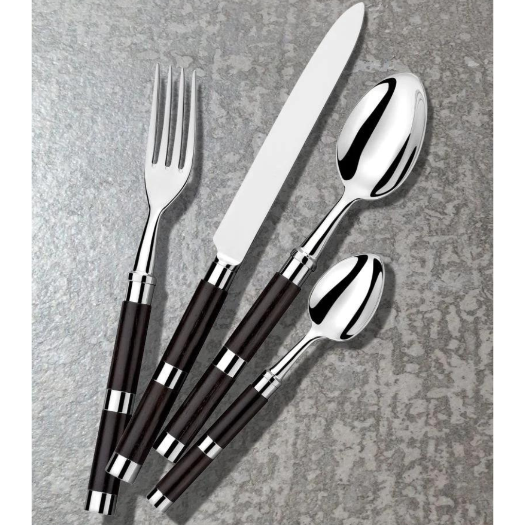 The Alain Saint-Joanis Carla Flatware Ebony 5 Piece Setting—hand-polished in France—features fork, knife, tablespoon, teaspoon, and salad fork with dark handles, arranged diagonally on a textured gray surface.