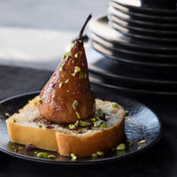 A caramelized pear rests on bread topped with pistachios, served on L'Objet Alchimie Dinnerware Black plate, with stacked black dishes from the same collection blurred in the background.