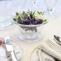 The William Yeoward Alice Bowl, a small handmade crystal piece featuring purple, white, and green flowers, sits on a white tablecloth beside plates and neatly wrapped cutlery.