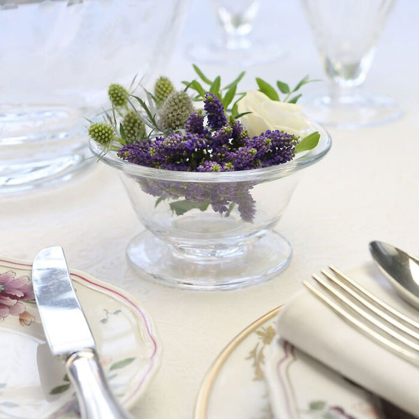 The William Yeoward Alice Bowl, a small handmade crystal piece featuring purple, white, and green flowers, sits on a white tablecloth beside plates and neatly wrapped cutlery.