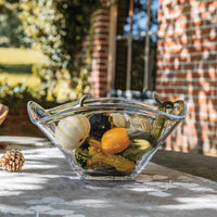 The Simon Pearce Woodbury Bowl, filled with assorted decorative gourds and squash, sits on an outdoor table with a blurred brick building and greenery in the background.