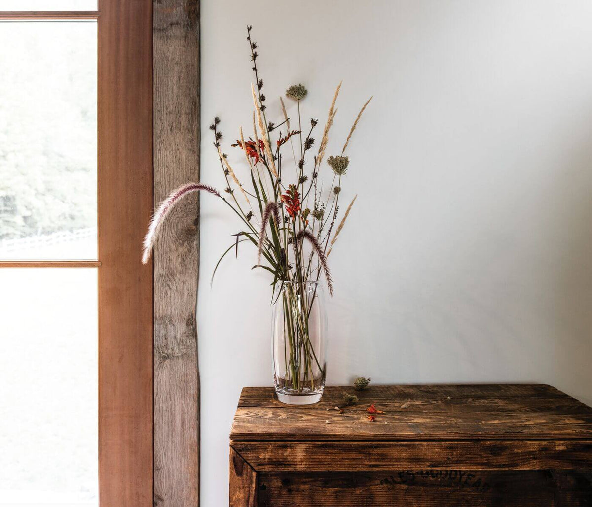 A Simon Pearce Addison Vase holds wildflowers and grasses on a rustic wooden chest, with natural light streaming in from a nearby window and a light-colored wall as the backdrop.