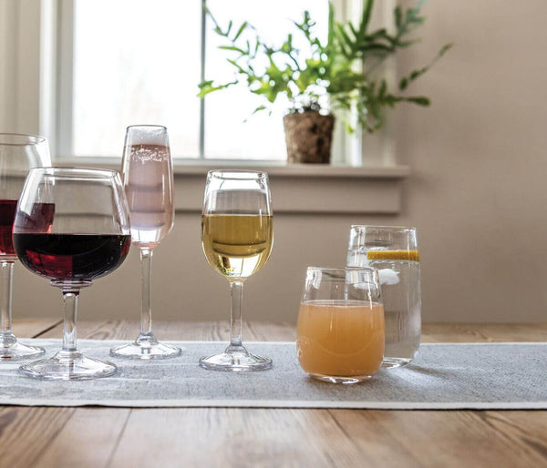 An assortment of drinks served in Simon Pearce Woodstock Tumblers—red wine, sparkling rosé, orange juice, and water with lemon—are set on a table with a light runner near a sunny window and potted plant. All tumblers are lead free.