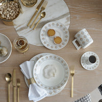 A modern table setting features Greg Natale’s Amalfi Dinnerware 3 Piece Set, gold utensils, coffee, a drink with dried citrus, two mini tarts, pistachios, and a white napkin on a light wood table.