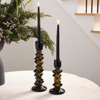 Two black tapered candles are lit in chunky Fortress Candleholders by Accent Decor, placed on a light surface with greenery and a staircase in the background.