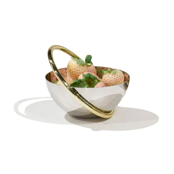 The Anna New York Ring Nut Bowl, crafted from shiny stainless steel and featuring a gold-colored handle, holds pale pink strawberries with green leaves against a white backdrop—ideal for luxurious home décor.