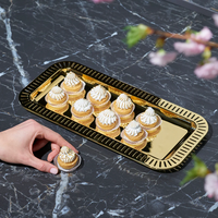 The Anna New York Striations Gold Small Rectangle Serving Tray holds ten mini desserts with whipped cream on a dark marble surface, while a hand reaches for a treat and pink flowers create a blurred foreground.