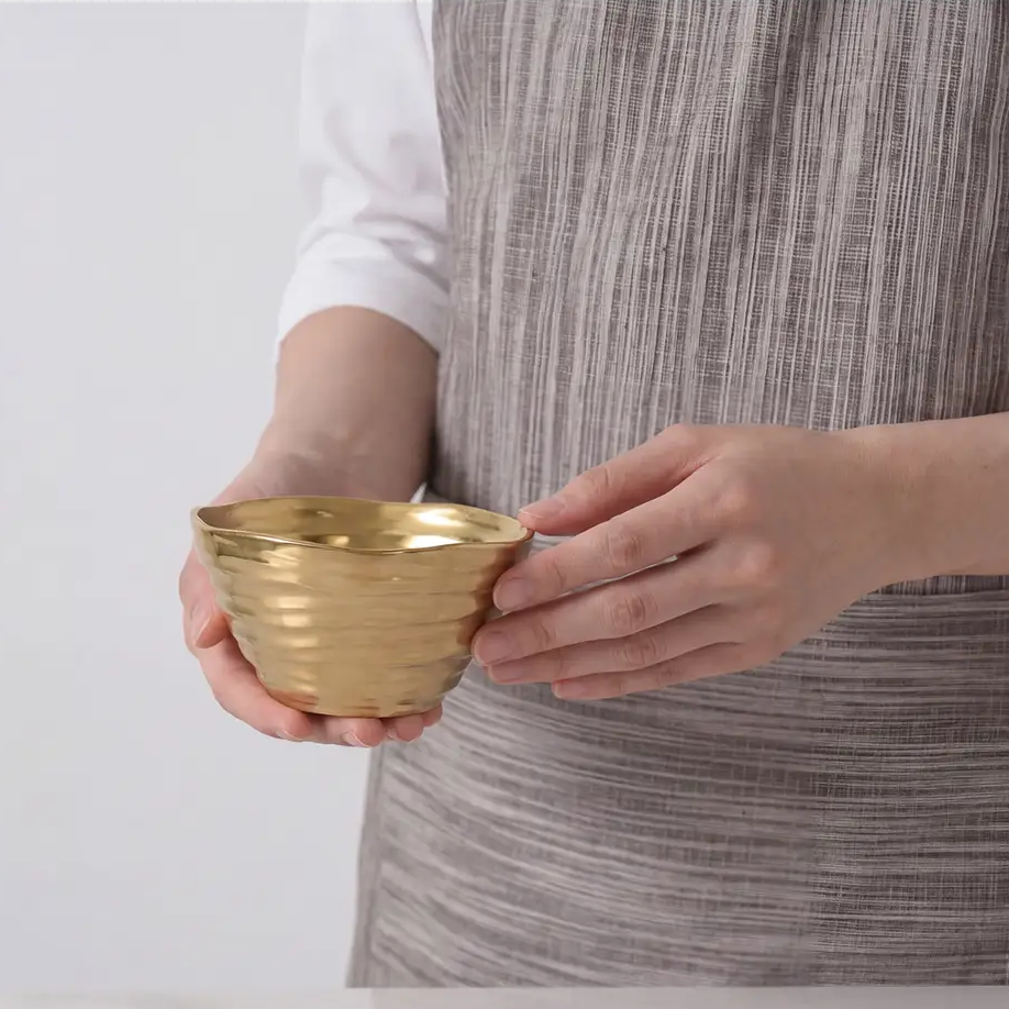 A person holds the Dorrato Gold Bowl - by PAMPA BAY with both hands, wearing a gray striped apron against a plain light background.