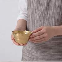 A person holds the Dorrato Gold Bowl - by PAMPA BAY with both hands, wearing a gray striped apron against a plain light background.