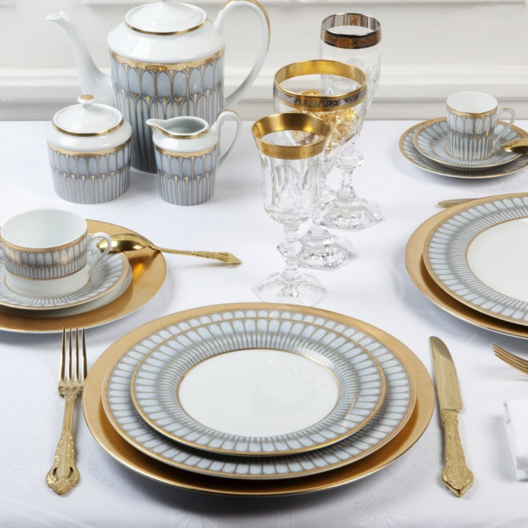 Elegant table setting with Arcades Gold Dinnerware by Philippe Deshoulieres, showcasing fine china adorned with gold and grey geometric patterns, crystal glasses, gold utensils, teacups, a teapot, creamer, and sugar bowl on a white tablecloth.