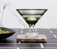A Footed Martini Glass by Tyler Barry Design, filled with a clear liquid, rests on a marble coaster beside green olives on black tiles—an elegant display of modern barware.