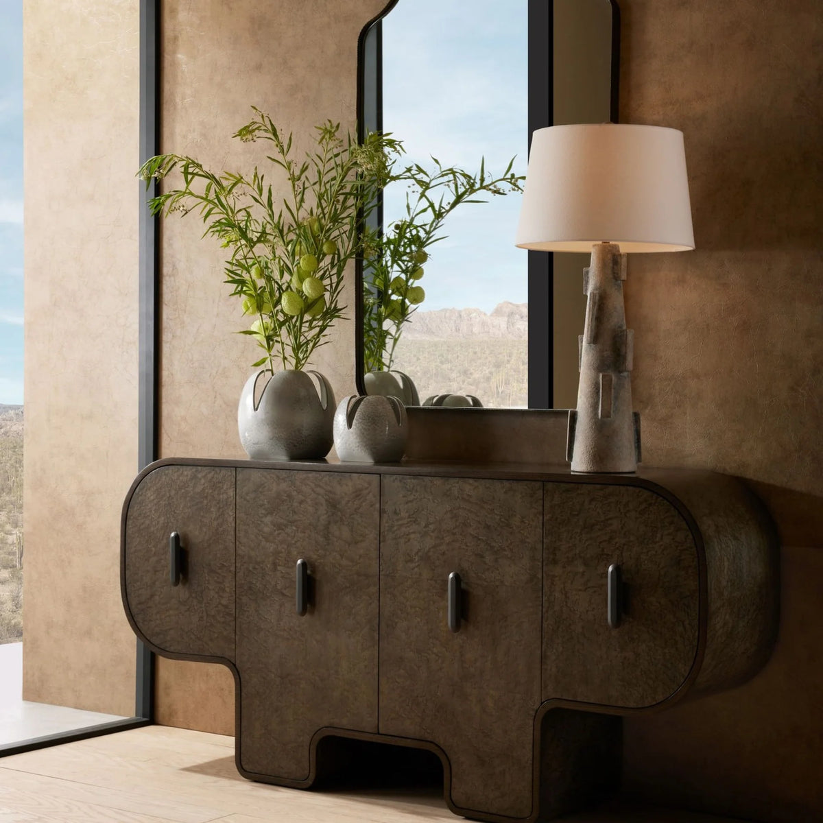 A modern sideboard with rounded edges displays the elegant Arteriors Amal Grey Glazed Vases Set of 2, filled with green branches, alongside a stacked stone lamp. A large mirror reflects the desert landscape visible through a tall window, while neutral-toned walls create a serene backdrop.