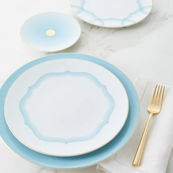 A gold fork lies on a white napkin beside Raynaud's Aura Dinnerware—white plates with blue geometric borders, gold trim, and scalloped edges—elegantly set on a white marble surface.