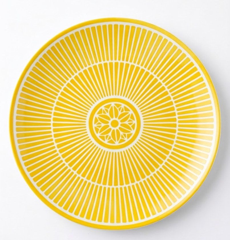 The Malmaison Riviera Dinnerware by Christofle features a round yellow plate with radiating geometric lines and a central decorative motif, inspired by French Riviera elegance.