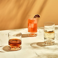 Three drinks served in the Nude Glassware Club Drinkware Set of 4—dark brown, tall pink cocktail with citrus, and clear bubbly with lemon—sit on a marble surface casting elegant shadows on a beige background.